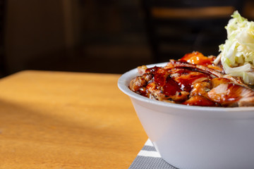 A view of a bowl of teriyaki chicken sitting on the right side of the frame, in a restaurant or kitchen setting.