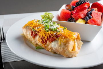 A view of a breakfast burrito plate with a bowl of fresh fruit, in a restaurant or kitchen setting.