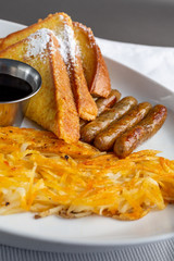 A view of a breakfast plate in a restaurant or kitchen setting, featuring hash browns, sausage links and French toast.