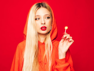 Portrait of young beautiful hipster bad girl in trendy red summer hoodie and earring in her nose.Sexy carefree smiling blond woman posing in studio.Positive model licking round sugar candy