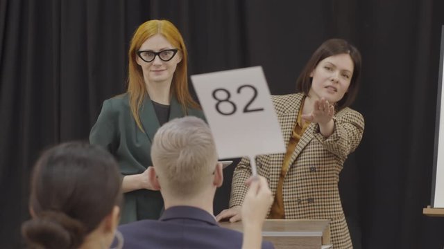 Beautiful female auctioneer standing at lectern beside colleague, pointing at paddles and calling bids during art auction