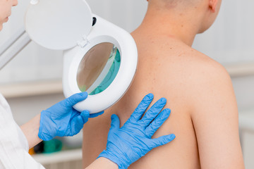 A dermatologist examines the skin on the body of a young man through a magnifying glass of a...