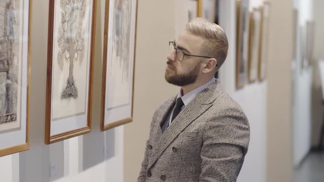 Handsome man in glasses and elegant suit standing in fine art gallery and examining drawing on the wall