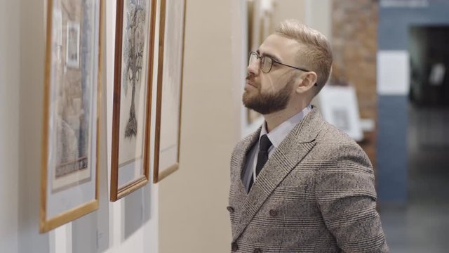 Intelligent caucasian man in glasses and elegant outfit standing in museum and examining artwork on the wall