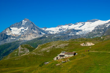 Fototapeta na wymiar During a sunny summer day with clear blue sky view of green pastures and traditional stone farmhouse and in the background snowy mountains in National Park Vanoise, French Alps.