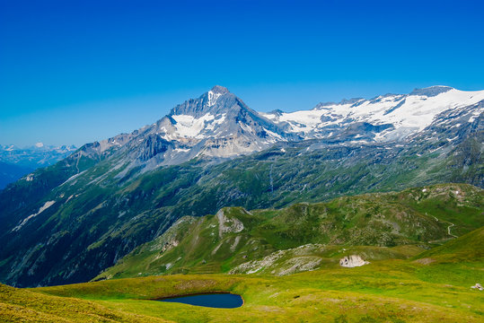 Beautiful summer alpine scenery, high mountain with snow on the slopes, green meadow with a small blue lake, hiking trails and clear blue sky in the background, French Alps, Vanoise National Park. 