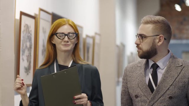 Attractive female gallery docent holding clipboard, walking and discussing modern drawing on the wall with male visitor while showing him collection of contemporary art