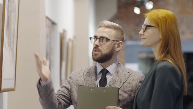 Male gallery guide holding clipboard, pointing at picture on the wall and discussing it with redhead woman while presenting modern art collection to her