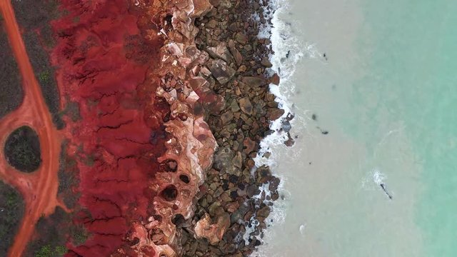 Aerial zoom out view over bright red sand and blue ocean along coastline of Broome, Western Australia