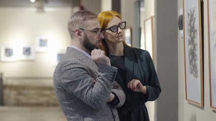 Professional male and female art critics standing in exhibition hall and discussing artwork on the wall while visiting gallery together