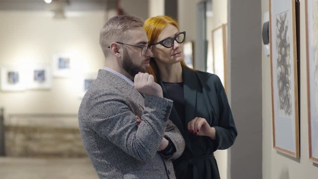 Professional male and female art critics standing in exhibition hall and discussing artwork on the wall while visiting gallery together