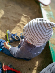 A kid sitting in a sandbox wearing a striped cap and playing with a rake