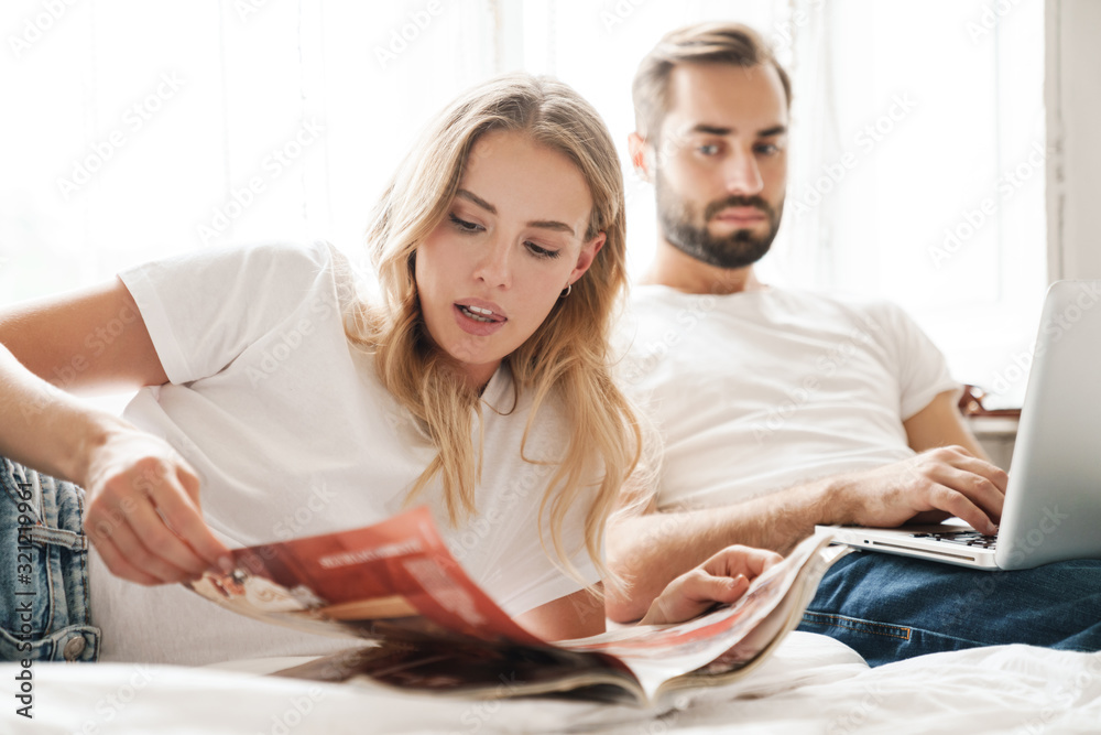 Sticker couple indoors at home reading magazine using laptop computer.