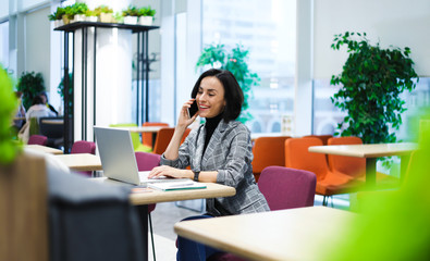 In the coworking. Side-view photo of a cheerful woman in smart casual clothes, talking on the phone...