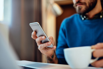 Close up of good-looking caucasian young businessman sitting in his office, holding cup of coffee and looking at smart phone. Selective focus on smart phone.