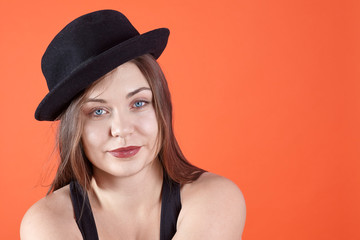 Young attractive caucasian brunette woman with bright blue eyes and dark lipstick, wearing black bowler hat, smiles looking right to the camera. Isolated on coral background, studio, copy space.