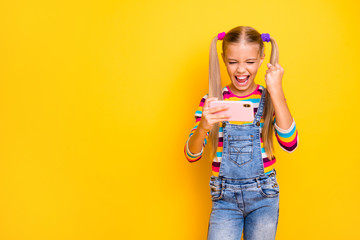 Portrait of ecstatic excited child girl use smartphone play online video games raise fists scream yeah wear striped sweater jumper denim jeans isolated over yellow bright color background