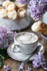 Cup of coffee and cake horns from puff pastry with vanilla cream in a metal box in spring still life with a bouquet of lilacs on a wooden table.