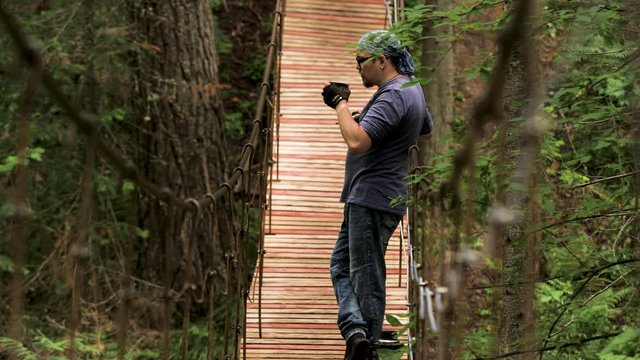 Side view of a man standing and having a rest on the wooden suspension bridge. Stock footage. Man looking into the distance and smoking a pipe in the forest.