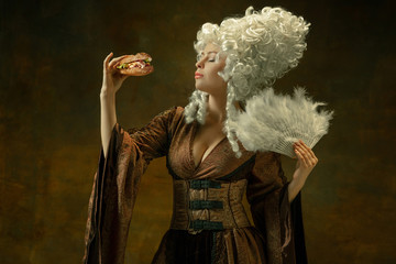Eating burger impressed. Portrait of medieval young woman in brown vintage clothing on dark background. Female model as a duchess, royal person. Concept of comparison of eras, modern, fashion, beauty.
