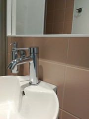 Hotel bathroom interior with round white sink and glossy metal details. Metal bathroom tap, soap dispenser and mirror in front of terra cotta tiles.