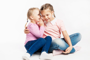 Two little girlfriends in jeans and pink sweaters gossip. White background.