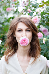 closeup vertical portrait of beautiful young woman brunette with pink rose bush on background. Girl dressed in a white blouse, looks at camera and holds a flower in her mouth