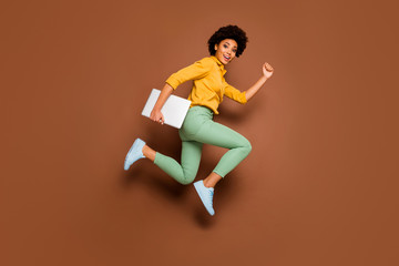 Fototapeta na wymiar Full length photo of funky dark skin lady jump high hold notebook hurry classes lessons schoolgirl wear yellow shirt green pants footwear isolated brown color background
