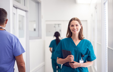 Portrait Of Smiling Female Doctor Wearing Scrubs In Busy Hospital Corridor Holding Digital Tablet