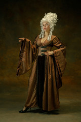 Elegance posing. Portrait of medieval young woman in brown vintage clothing on dark background. Female model as a duchess, royal person. Concept of comparison of eras, modern, fashion, beauty.