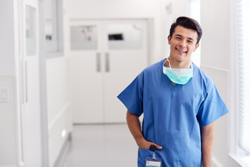 Portrait Of Young Male Doctor Wearing Scrubs Standing In Hospital Corridor - Powered by Adobe