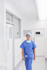 Young Male Doctor Wearing Scrubs Walking Along Hospital Corridor