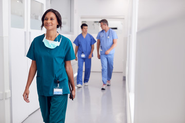 Female Doctor Wearing Scrubs In Hospital Walking Along Corridor Holding Digital Tablet