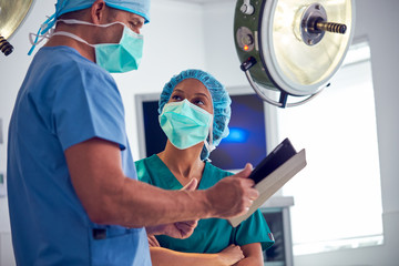 Male And Female Surgeons Wearing Scrubs Looking At Digital Tablet In Hospital Operating Theater