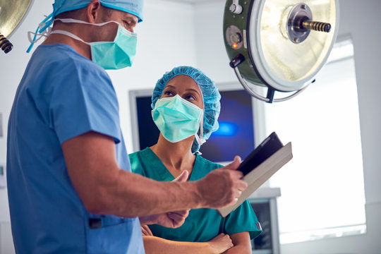 Male And Female Surgeons Wearing Scrubs Looking At Digital Tablet In Hospital Operating Theater - Powered by Adobe