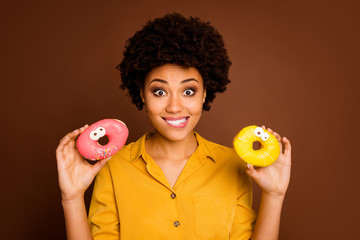 Photo of pretty dark skin lady hold two colorful donuts caramel eyes human faces painting carefree desire hungry bite lips wear yellow shirt isolated brown color background