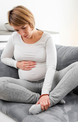 Fototapeta premium Pregnant tired exhausted woman with stomach, leg and pain issues at home on a couch, being sick.