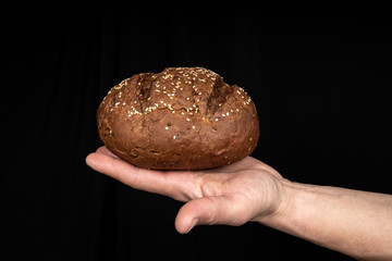 Man's hands hold tasty fresh loaf of dark bread with sesame seeds on black background