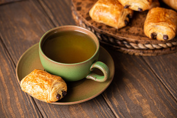 Green cup of tea with mini chocolate bun, puff pastry on old wooden table. Tasty tea break concept, copy space.