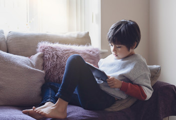 Candid shot cute little boy watching cartoons on tablet, Portrait of School kid sitting on sofa with serious face playing games on smart phone. Warm and cozy scene in  Pastel tone