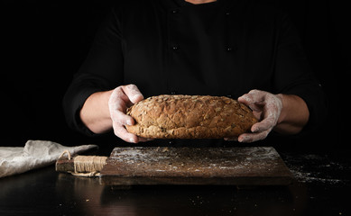 male hands are holding brown baked rye bread over wooden board with flour