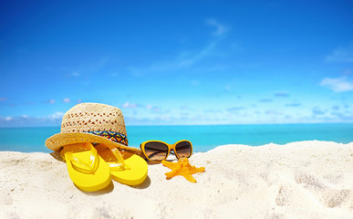 Concept summer beach holiday. Beach accessories - straw hat, glasses, starfish, yellow flip-flops on sandy tropical beach against blue sky with clouds on bright sunny day. Beautiful colorful image.