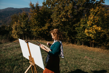 Young female artist working on painting outdoors, in the autumn landscape. She is in front of the canvas and drawing.She holds oil paints, artist brushes, canvas and palette. 