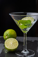 Close-up of cocktail glass with mint leaves and slice of lime on black background with a lime and a half with selective focus on vertical