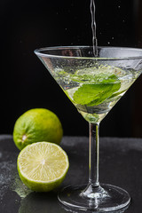 Close-up of cocktail glass with mint leaves and lime slice, pouring water, with splashes, on black background with a lime and a half with selective focus on vertical
