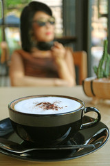 Closeup cappuccino in a black cup with blurry woman drinking coffee in the backdrop