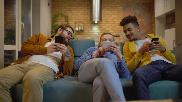 Male friends sitting on couch with gadgets, talking having fun with mobile apps