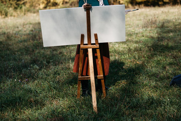 Young female artist working on painting outdoors, in the autumn landscape. She is in front of the canvas and drawing.She holds oil paints, artist brushes, canvas and palette. 