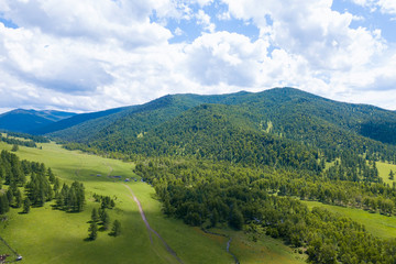 Aerial landscape with mountains, green trees, field, road and river under blue sky and clouds in summer