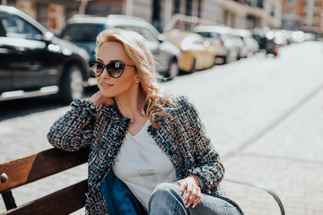 stylish young woman on a bench resting in city. girl wears sunglasses and a jacket in a cage on the background of new houses 